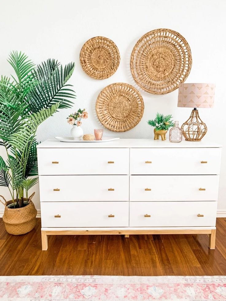 a white dresser topped with lots of drawers next to a potted plant and two wicker wall hangings