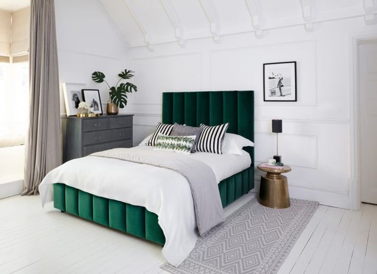 a bed with green velvet headboard and pillows in a white walled room, next to a window