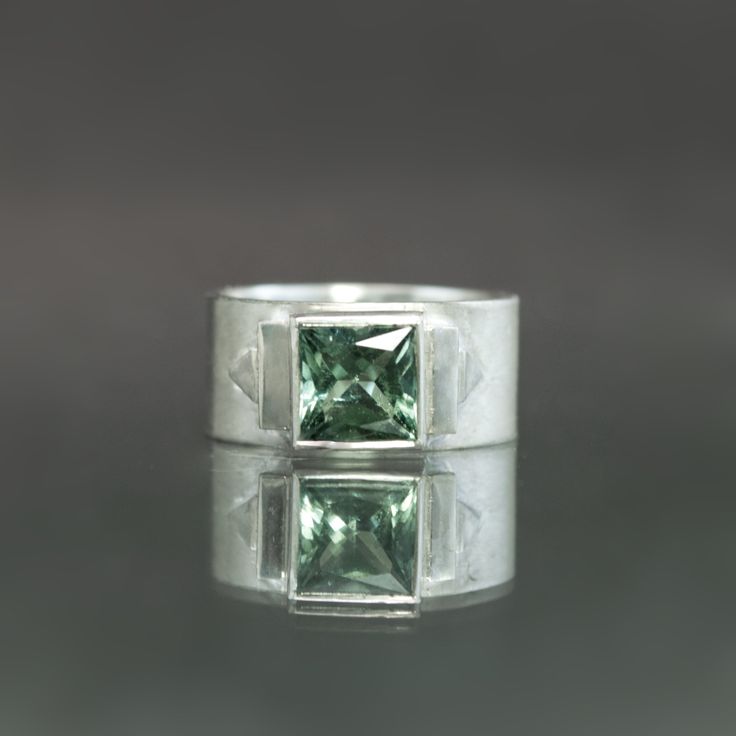 Specifications: Metal: Sterling Silver Stone: Natural Green Tourmaline Square, 1.85cts Size: US 7 Entirely hand-fabricated in sterling silver, this wide band ring presents a 1.85-carat deep moss hued tourmaline in glorious Art Deco form. A subtle contrast of matte and polished finishes flank the natural gem on either side, placed in a geometric arrangement inspired by Jazz Age architectural details. Sized to a 7, this ring is one of kind by nature of its stone and available to ship within 3 busi Modern Emerald Ring With Center Stone, Modern Sterling Silver Emerald Cut Sapphire Ring, Silver Emerald Cut Tourmaline Jewelry, Modern Silver Ring With Emerald Center Stone, Modern Silver Emerald Ring With Center Stone, Modern Sterling Silver Jewelry With Emerald Cut, Modern Sterling Silver Jewelry With Accent Stones, Modern Tourmaline Wedding Rings, Polished Tourmaline Jewelry For Anniversary