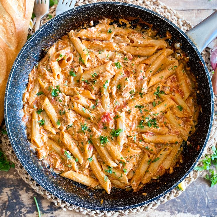 a skillet filled with pasta and sauce on top of a table next to bread