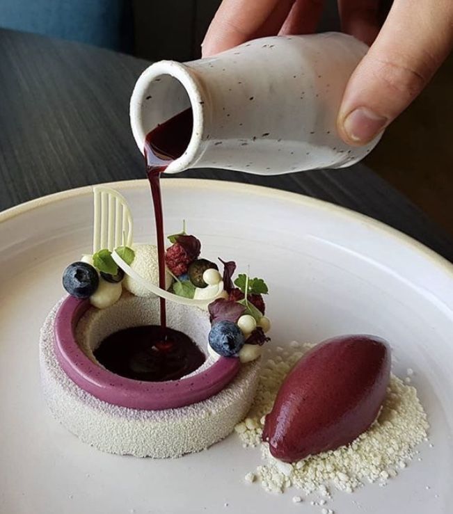 a person pouring sauce onto a dessert on a white plate with berries and blueberries