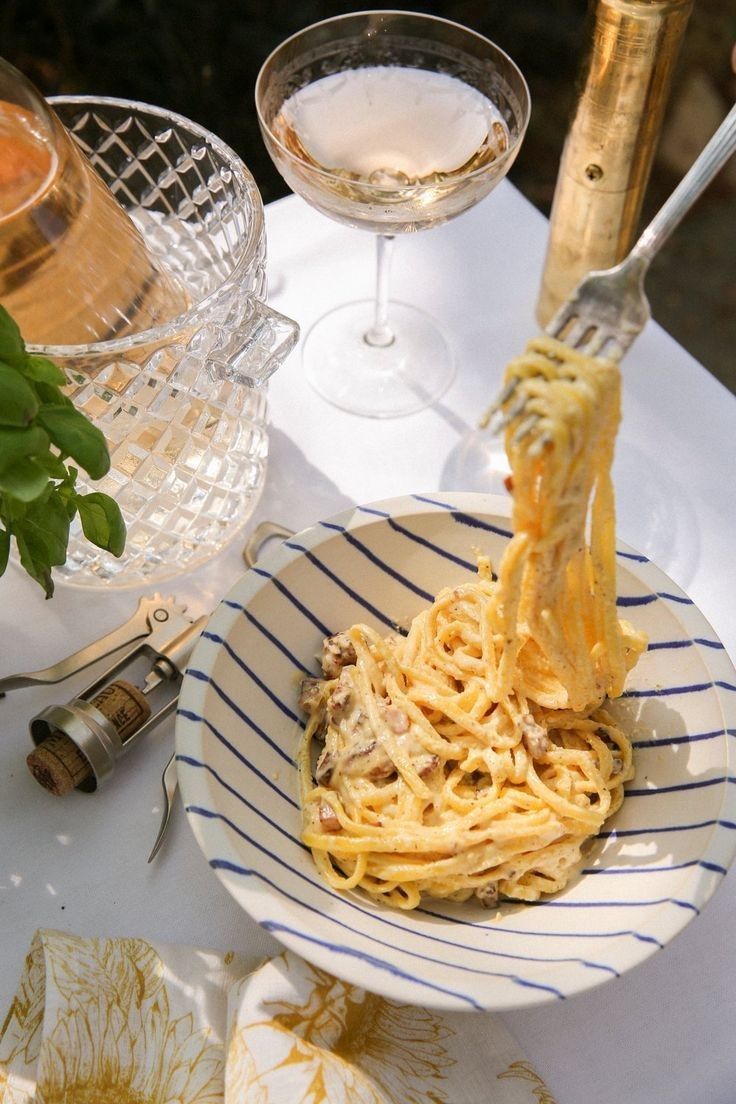 a plate of spaghetti being lifted from a bowl with wine glasses and utensils in the background