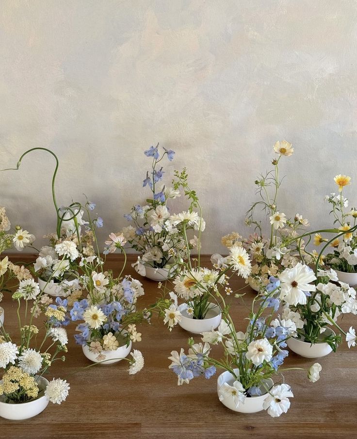 several white vases filled with different types of flowers on a wooden table in front of a wall