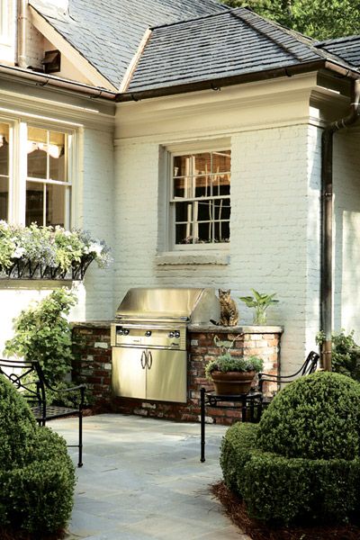 an outdoor kitchen is shown in front of a white house with bushes and shrubs around it