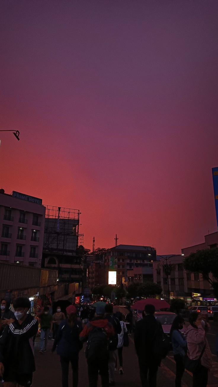 the sky is pink and purple as people are walking down the street at night with buildings in the background