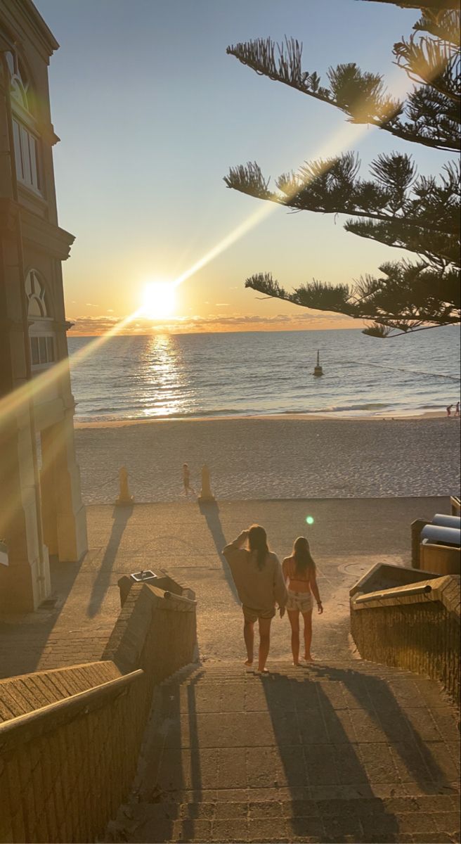 two girls walking down the sidewalk towards the beach at sunset, with the sun setting in the background