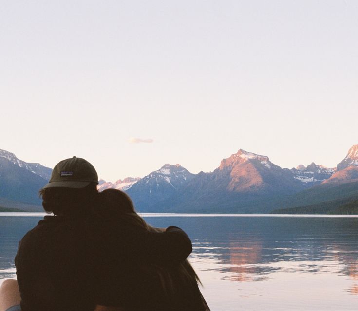 two people sitting on the edge of a body of water with mountains in the background