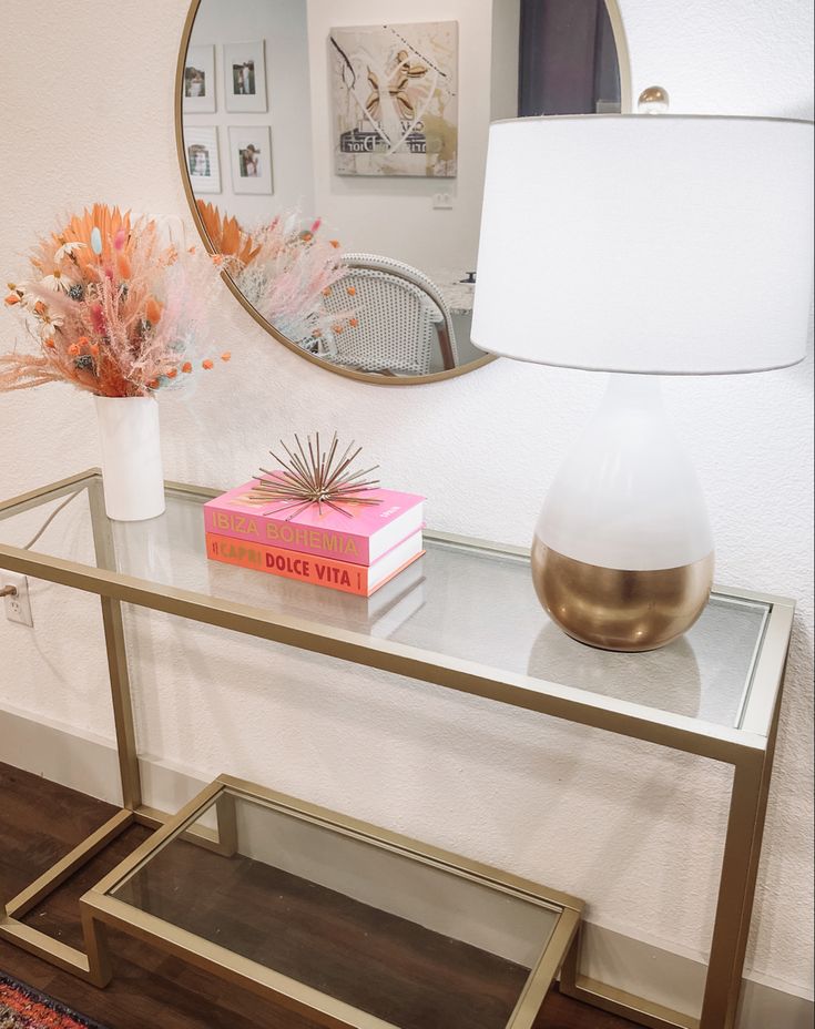 a table with a mirror, vase and books on it in front of a lamp