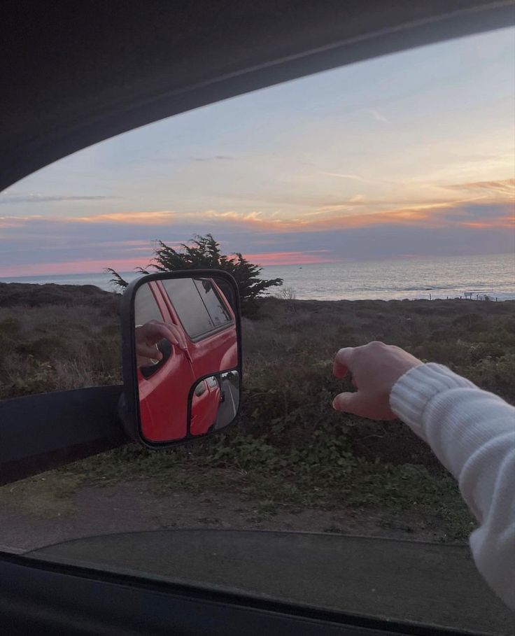 a person's reflection in the side view mirror of a red car next to an ocean