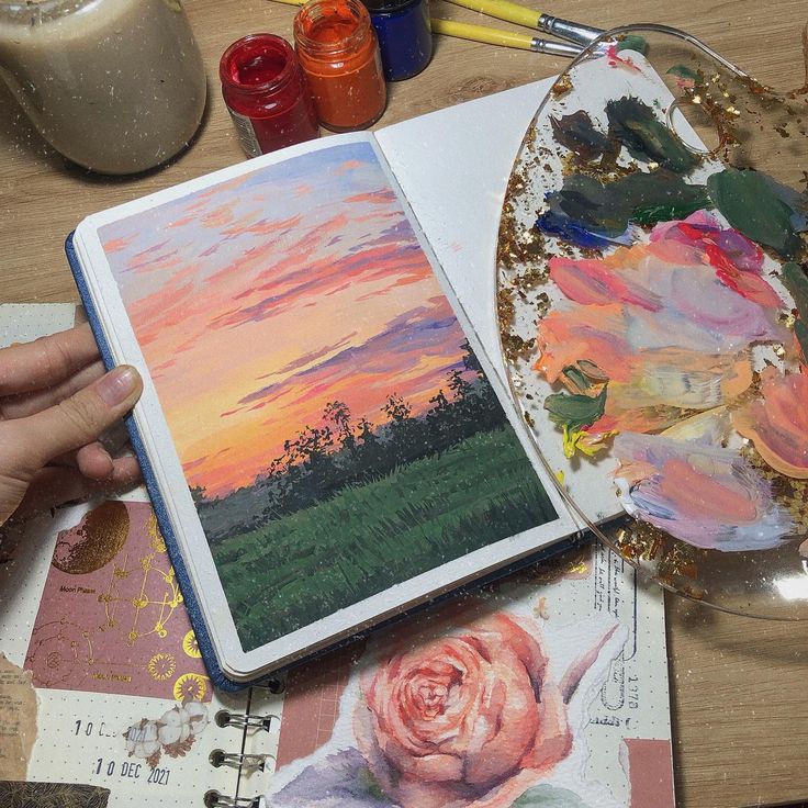 a person holding an open book on top of a table next to some art supplies