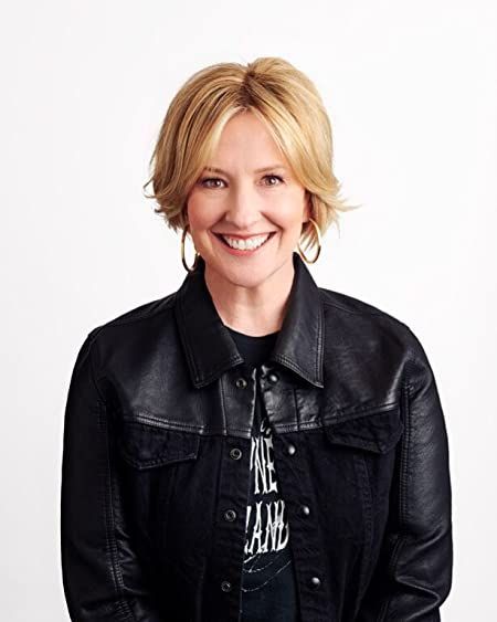 a woman with blonde hair wearing a black jacket and smiling at the camera while standing in front of a white background