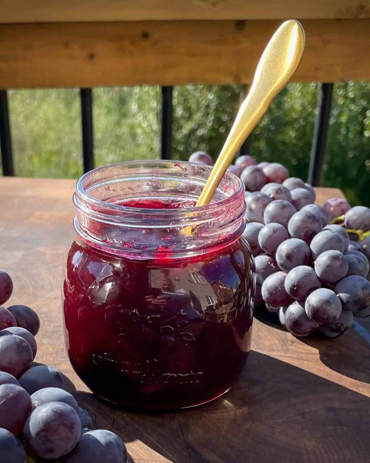 a glass jar filled with red wine next to grapes and a wooden spoon on a table