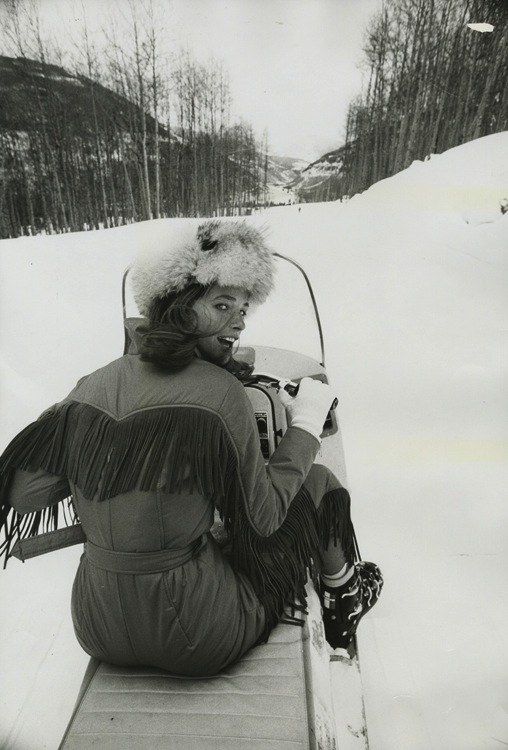 a woman sitting in the snow with her skis on and wearing a fur hat