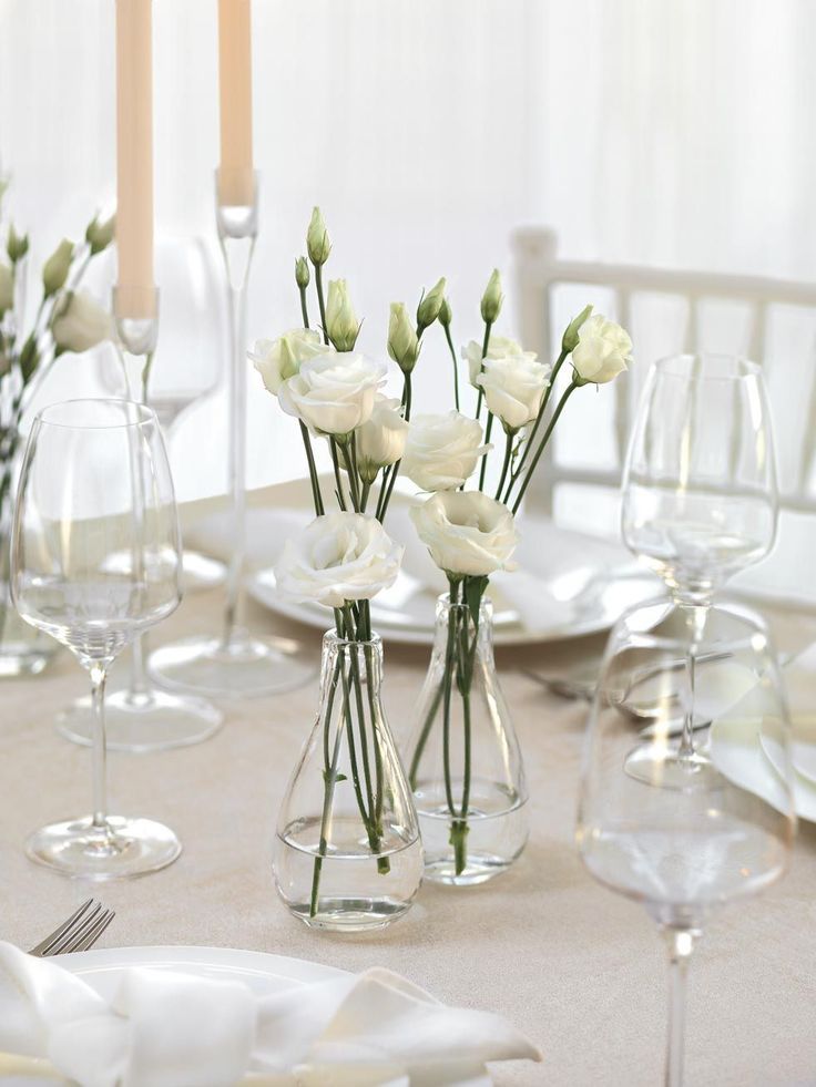 white flowers in vases sitting on a table with wine glasses and candles behind them