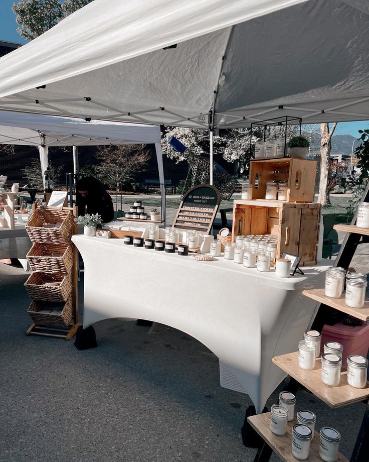 an outdoor market with white tables covered in jars and containers under a tented area