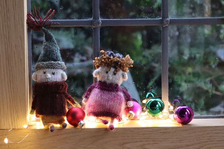 two knitted gnomes sitting on a window sill with christmas ornaments around them