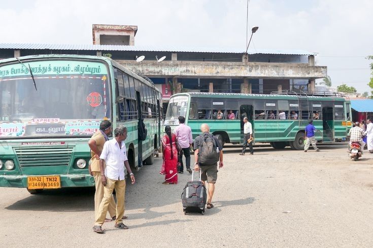 several people are walking towards two green buses on the street, one is carrying luggage