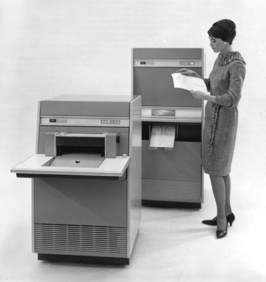 a woman standing next to two old computer machines with papers in front of her and another machine behind her