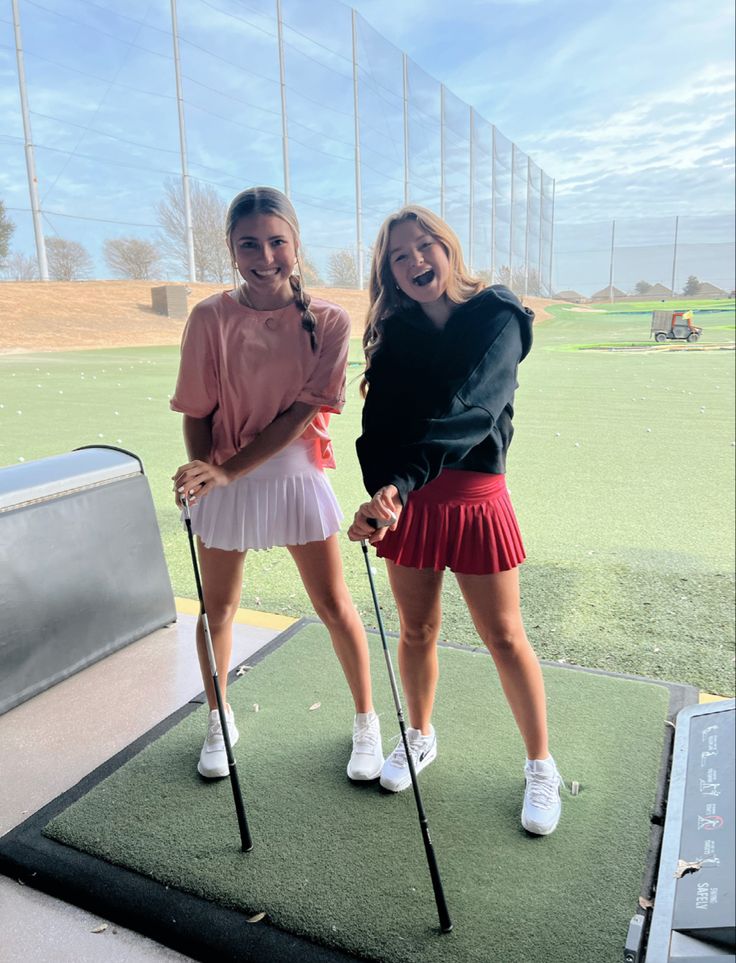 two women standing on top of a green field holding golf club's driver's poles