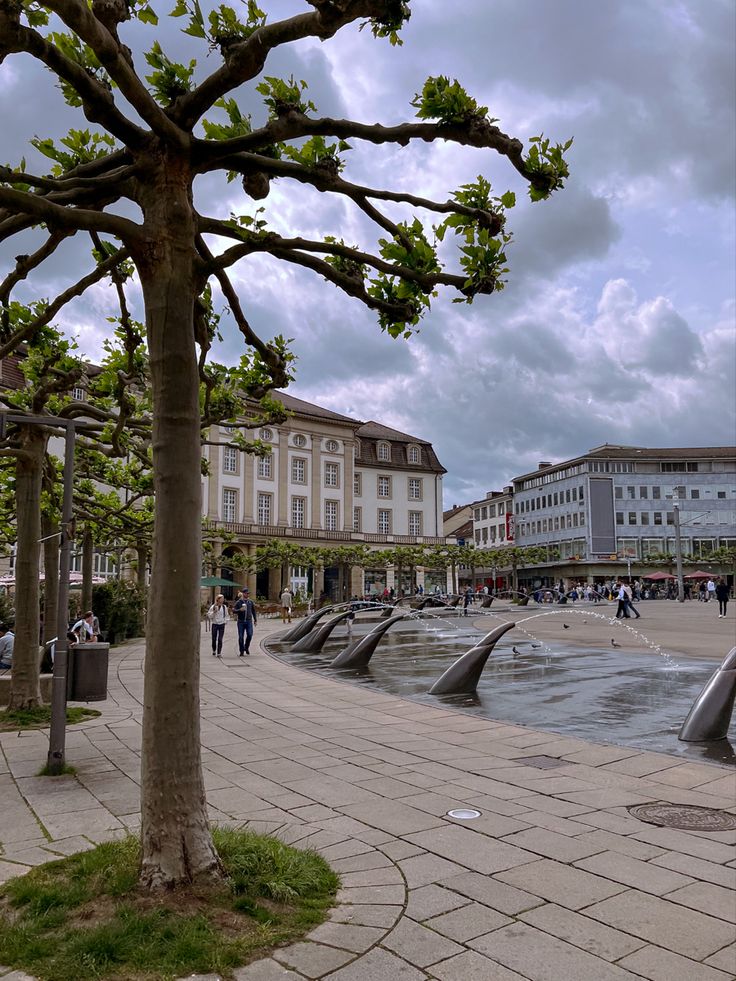 people are walking around in the city square with water fountains and trees on either side