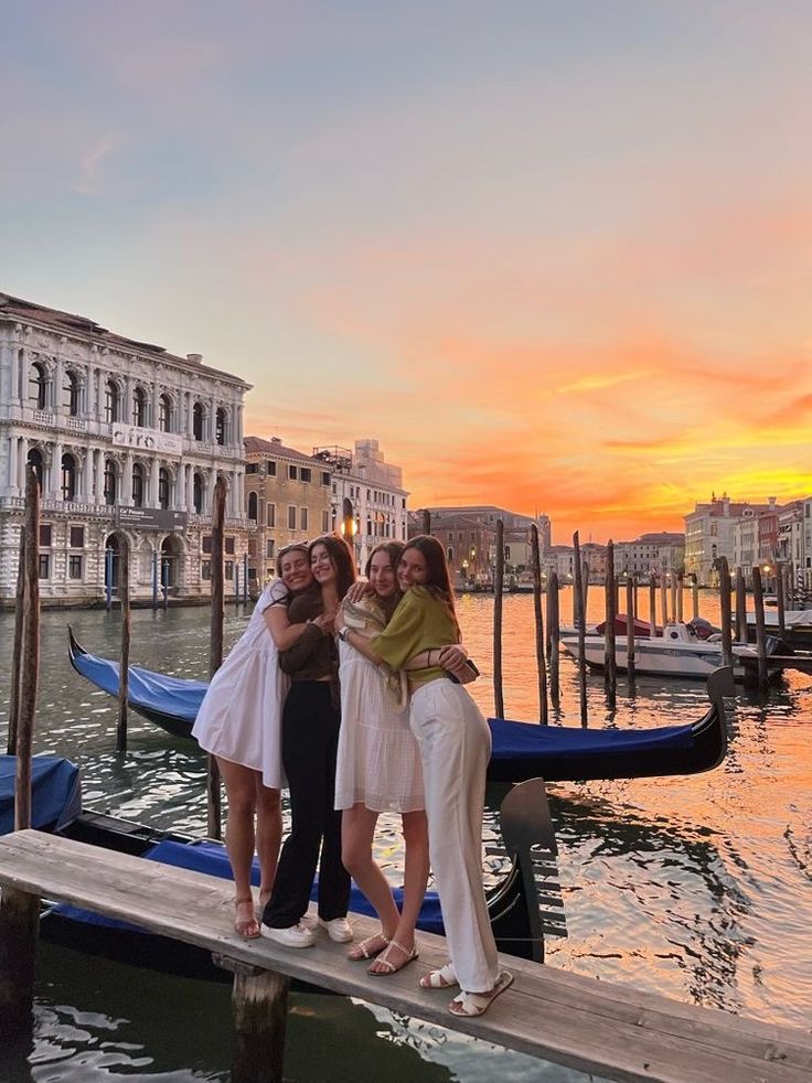 three women are standing on a dock in front of some gondolas at sunset