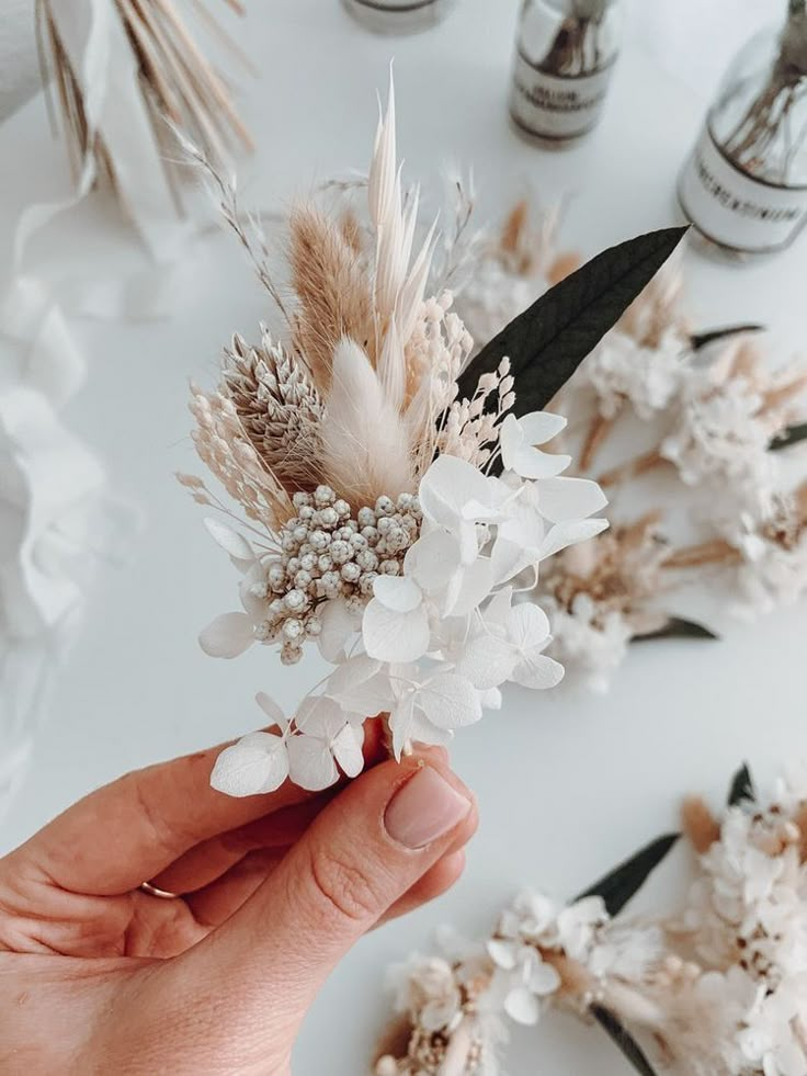 a person holding flowers in their hand on a white table with other items behind them