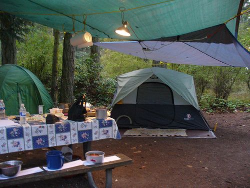 a tent set up in the woods with picnic table and chairs under it, next to a campfire