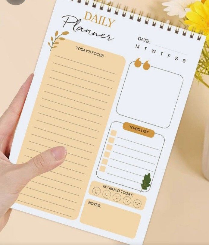 a hand holding a daily planner on top of a desk next to a vase with flowers