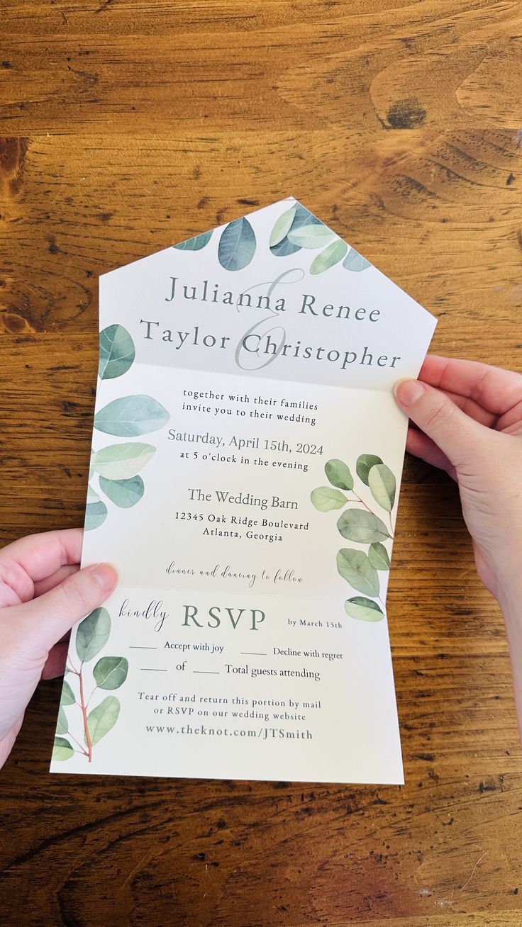 a person holding up a wedding card on top of a wooden table with greenery