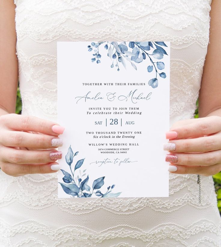 a woman holding up a wedding card in front of some flowers and greenery with her hands