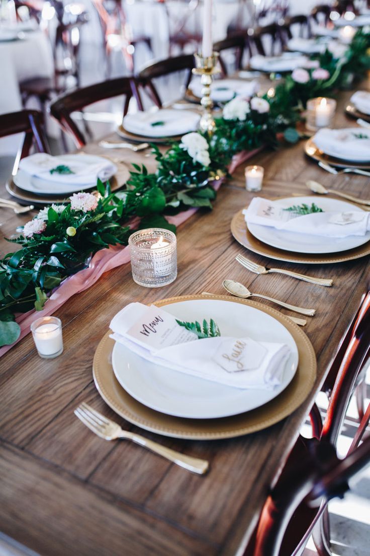 the table is set with white plates and place settings, greenery on each plate