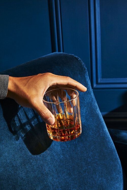 a hand holding a glass filled with liquid on top of a blue chair in front of a wall