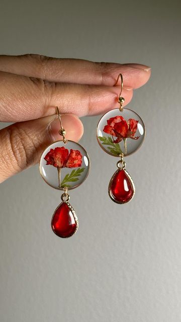 a woman's hand holding two red earrings with flowers on them and one dangling from it