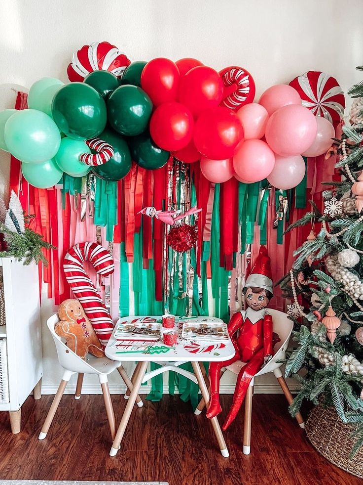 an elf is sitting at a table in front of a christmas tree with balloons and candy canes