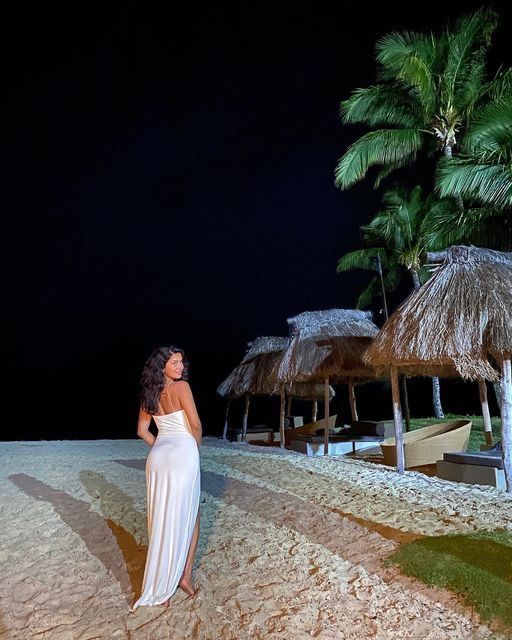 a woman in a white dress standing on the beach at night with palm trees and thatched umbrellas