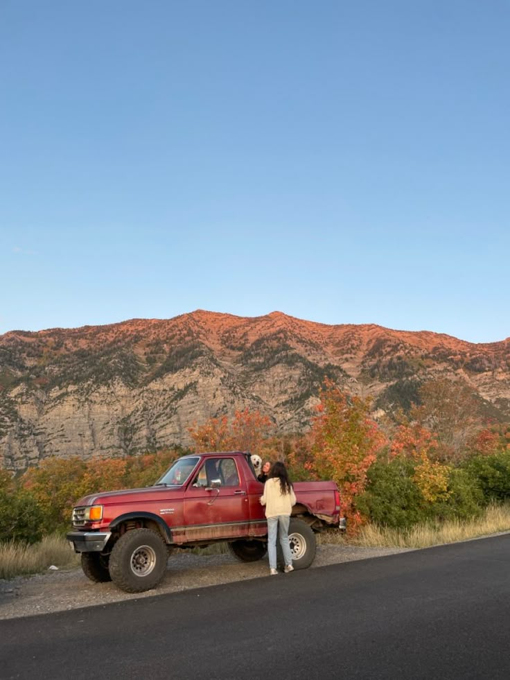 a woman standing next to a red truck on the side of a road with mountains in the background