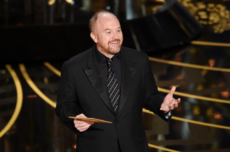 an older man in a black suit and tie holds his hand out while standing on stage