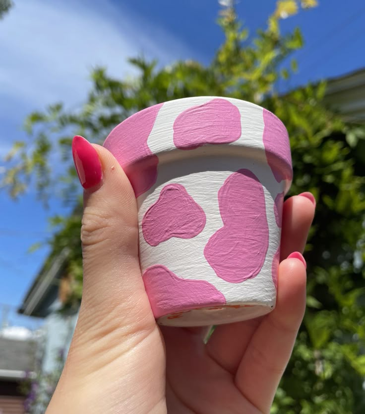 a hand holding a pink and white cup with hearts painted on the outside, in front of some trees