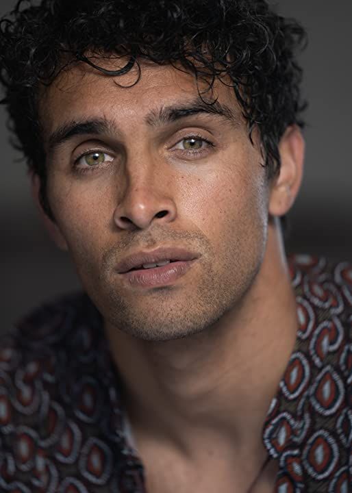 a man with curly hair looks at the camera while wearing a patterned shirt and looking off into the distance