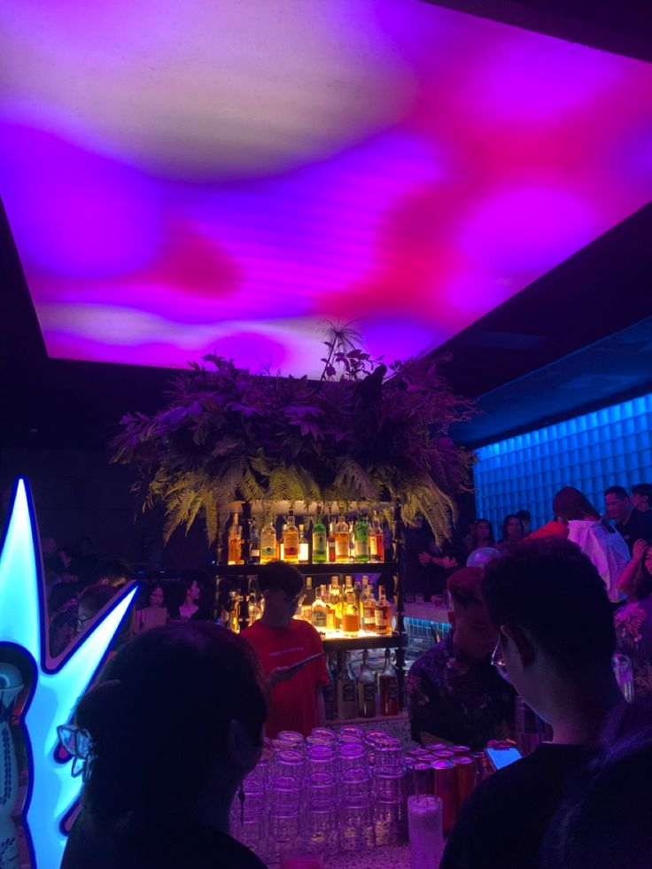 people are sitting at a bar with colorful lights on the ceiling and plants in vases