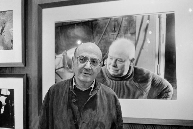 black and white photograph of man standing in front of art work on wall with multiple frames