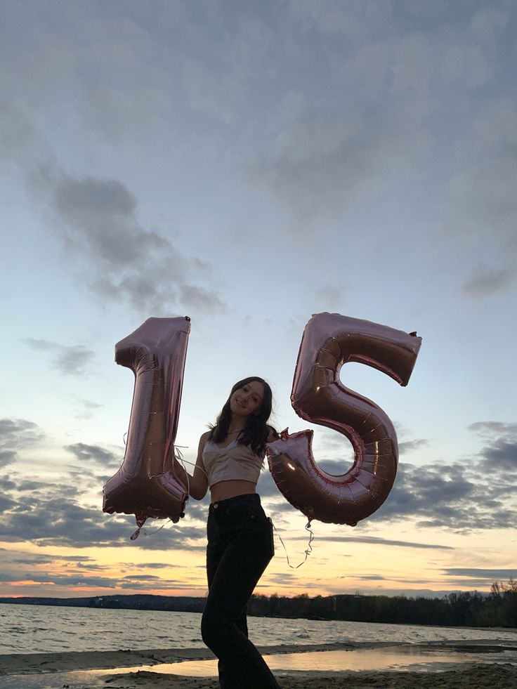 a woman standing on top of a beach holding two large balloons in the shape of the number fifteen