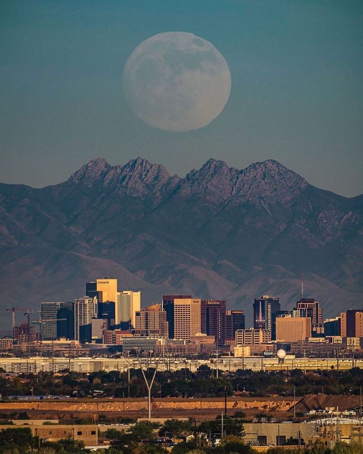 the full moon is setting over a city with mountains in the background