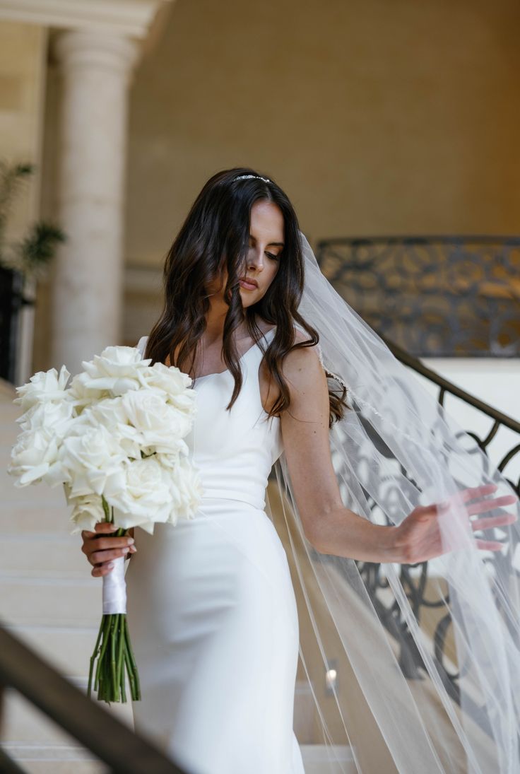 the bride is walking down the stairs with her veil blowing in the wind and holding a bouquet of white flowers