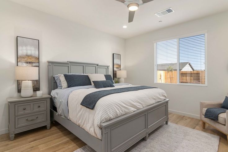 a bedroom with white walls and wood flooring has a large bed, two chairs, and a ceiling fan