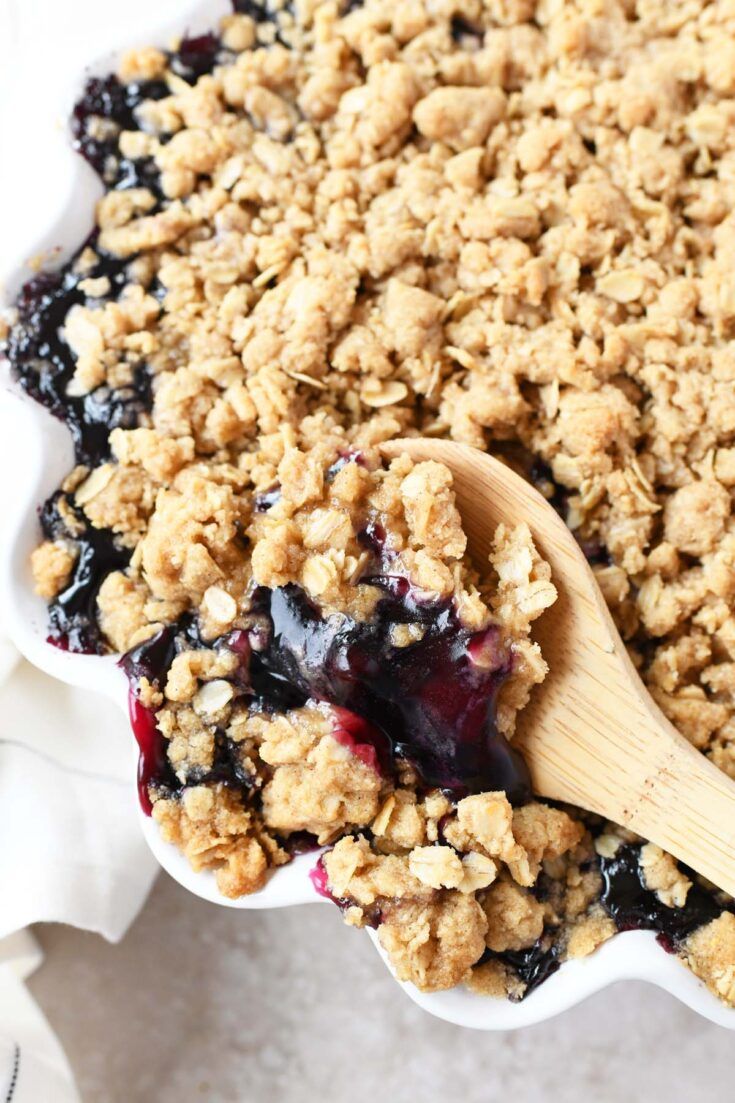 a blueberry crumbled dessert in a white dish with a wooden spoon on top