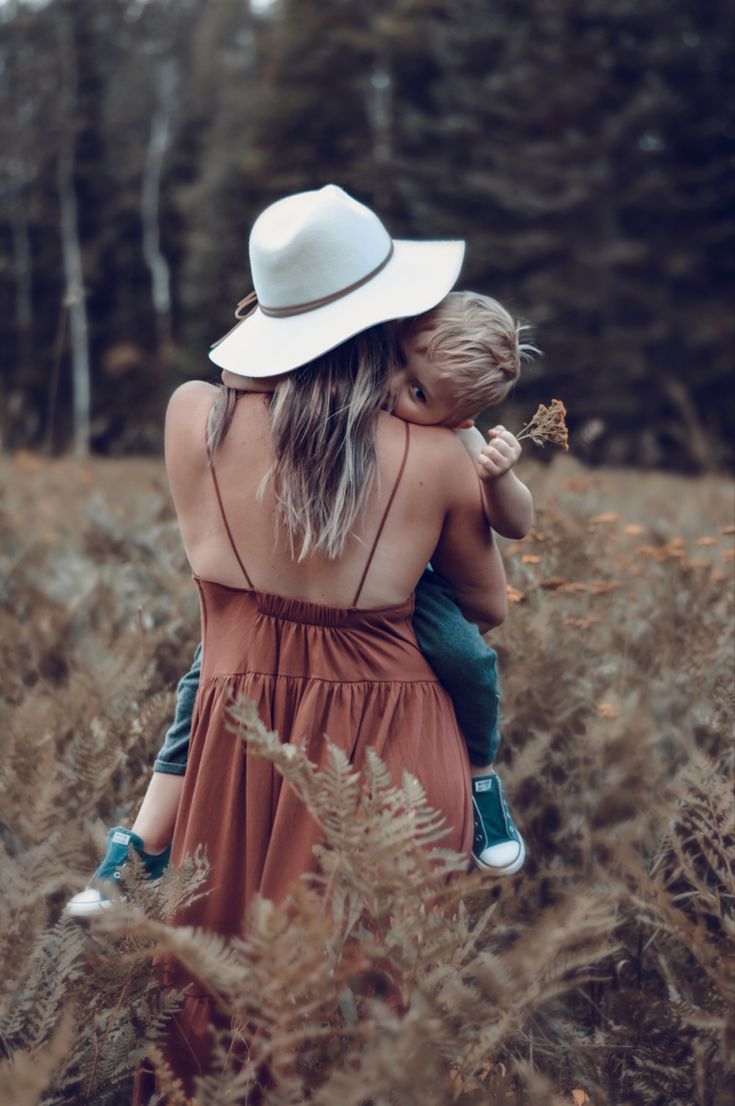 a woman in a white hat and dress holding a child while walking through tall grass