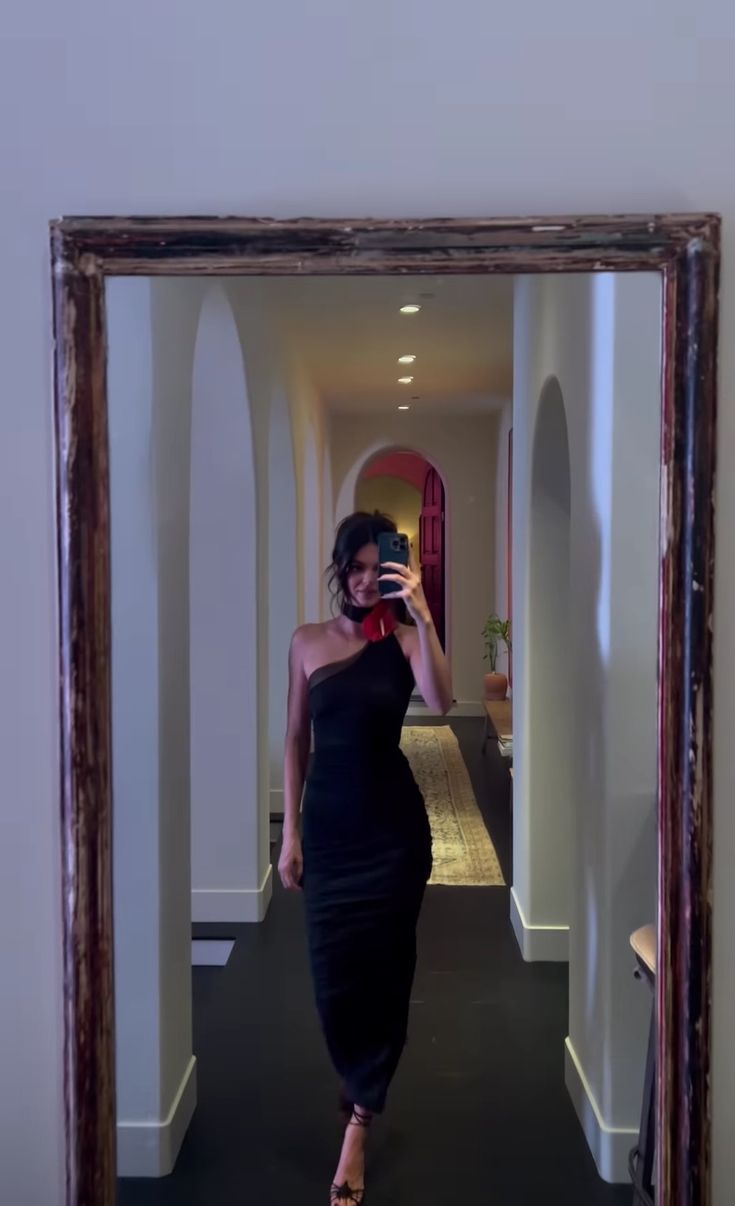 a woman in a black dress taking a selfie with her cell phone while walking down a hallway