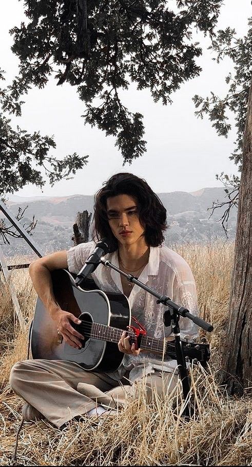 a man sitting in the grass playing a guitar and singing into a microphone with a tree behind him