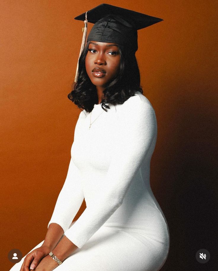 a woman wearing a graduation cap and gown sitting on a stool in front of an orange wall